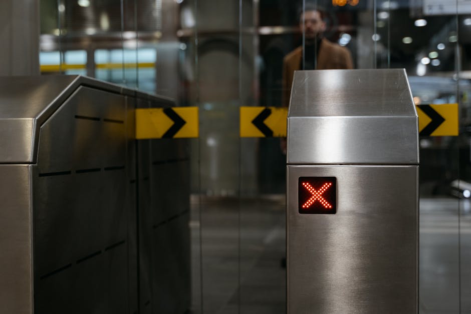 A close-up view of a metallic security barrier at an airport or train station entrance, with a bright red 'X' illuminated on the digital display panel indicating the gate is closed or inaccessible. Be